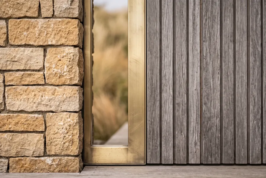 Limestone coursing, silvered timber battens, and a brass door reveal.